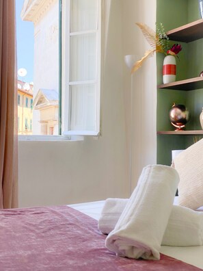 A hotel room with a bed, a window with white shutters, a green wall, and a shelf with decorative items. - OROROSA Lucca City Center Apartment (Lucca)