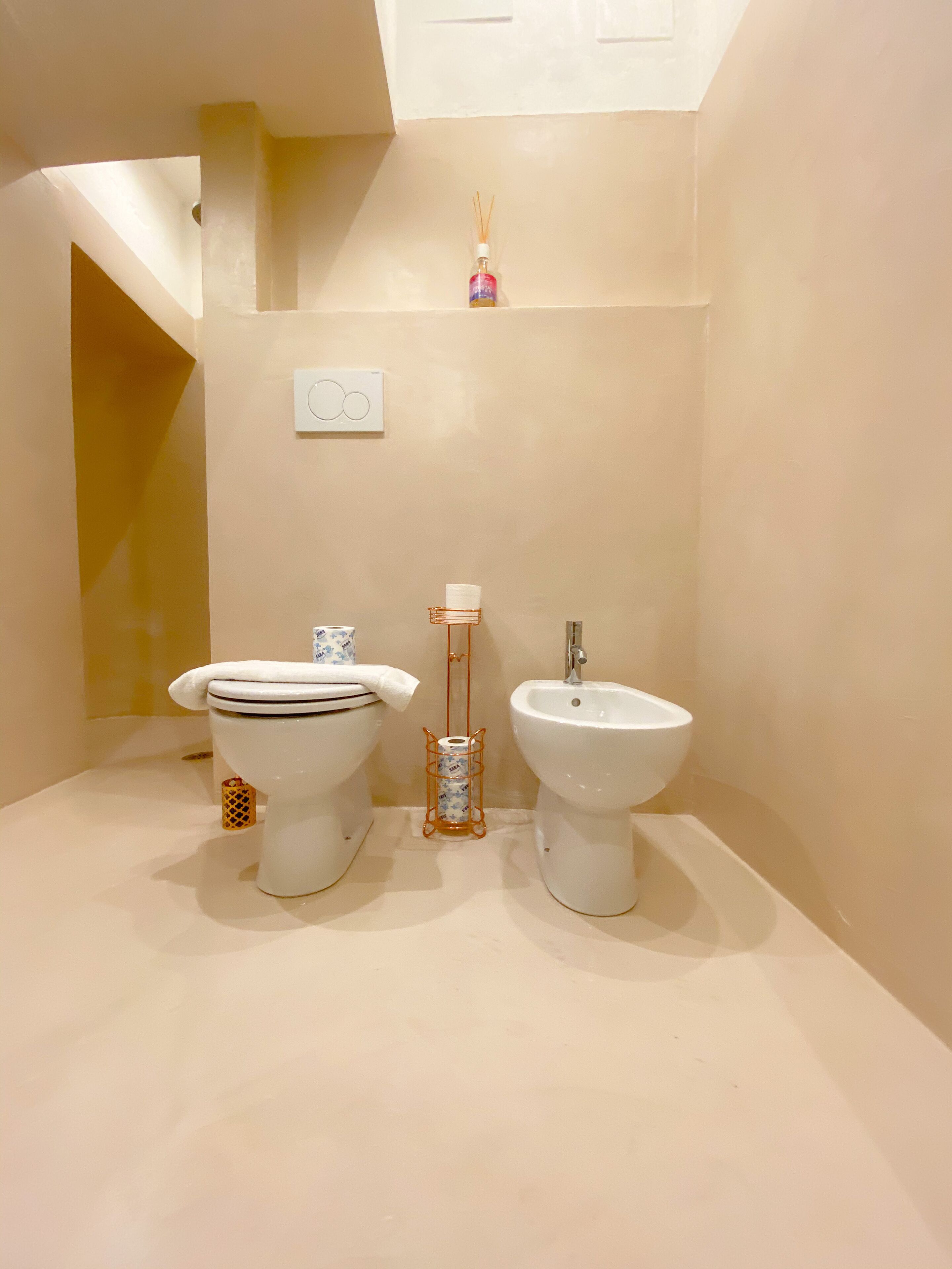 A bathroom with a toilet, bidet, and a small shelf with a bottle of air freshener.