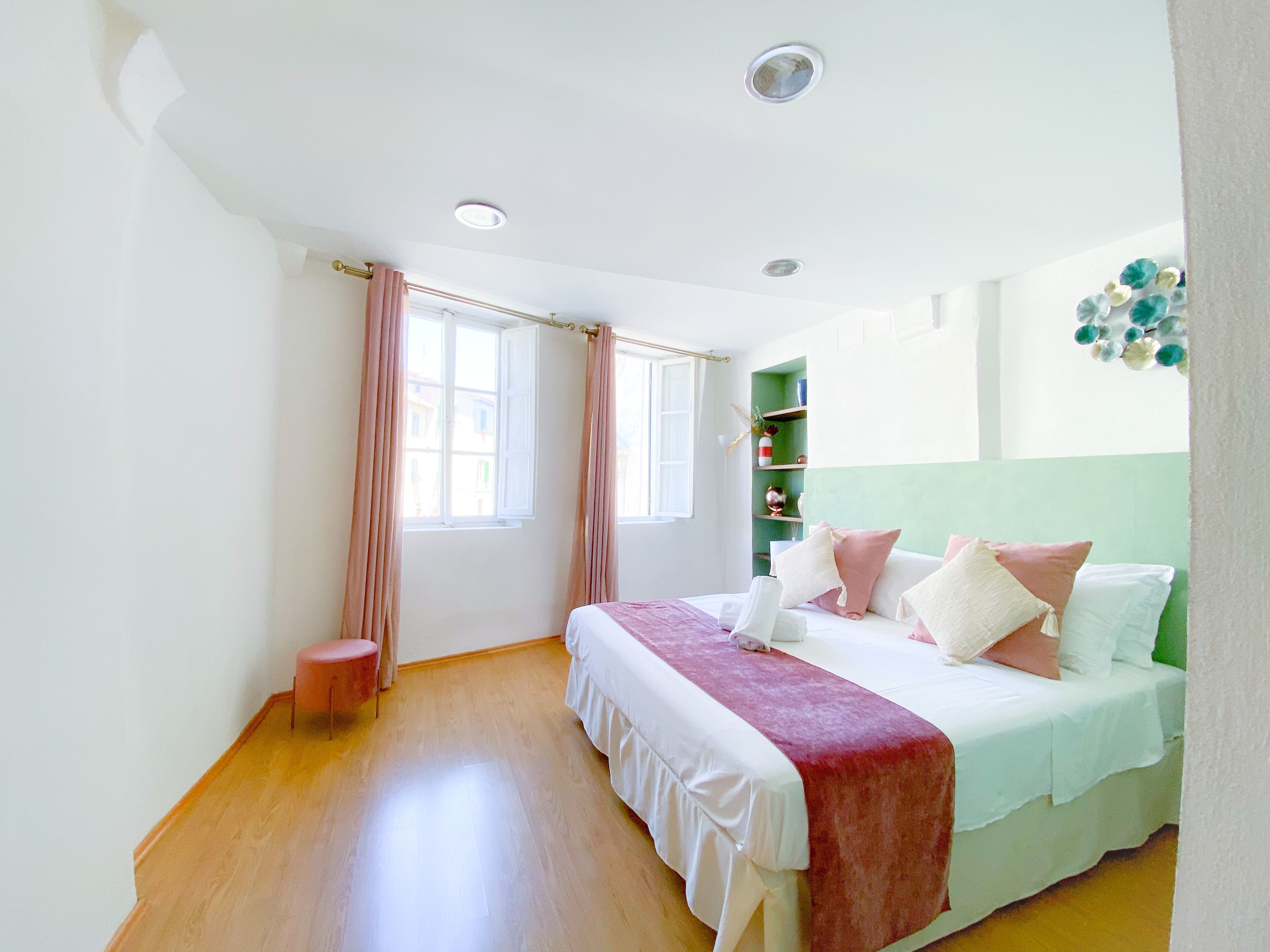 A bedroom with a bed, a small stool, a window with curtains, and a shelf with decorative items.