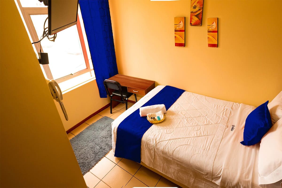 Habitación de hotel con cama, escritorio y silla. Se ve una ventana con cortinas, un teléfono y una obra de arte en la pared.