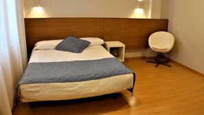 Un lit bien fait, avec une couverture bleue et des oreillers, une table de chevet, une chaise et des lampes fixées au mur.