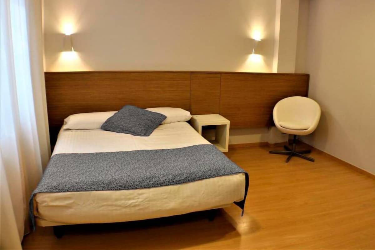 Una cama bien tendida con una manta azul y almohadas, una mesita de noche, una silla y luces fijadas a la pared.