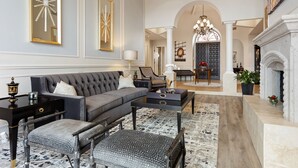 Un salón amplio con un sofá gris, un sillón estampado y una mesa de centro. Se observa una chimenea, un candelabro y un comedor al fondo.