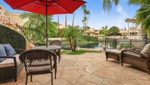 Un patio al aire libre con mobiliario de mimbre, una sombrilla roja y vistas a una piscina y palmeras.