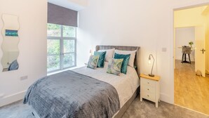 A bedroom with a large bed, a bedside table, a nightstand, a window with a view of greenery, and a door leading to another room. - 1 bedroom accommodation in Haworth (Haworth)