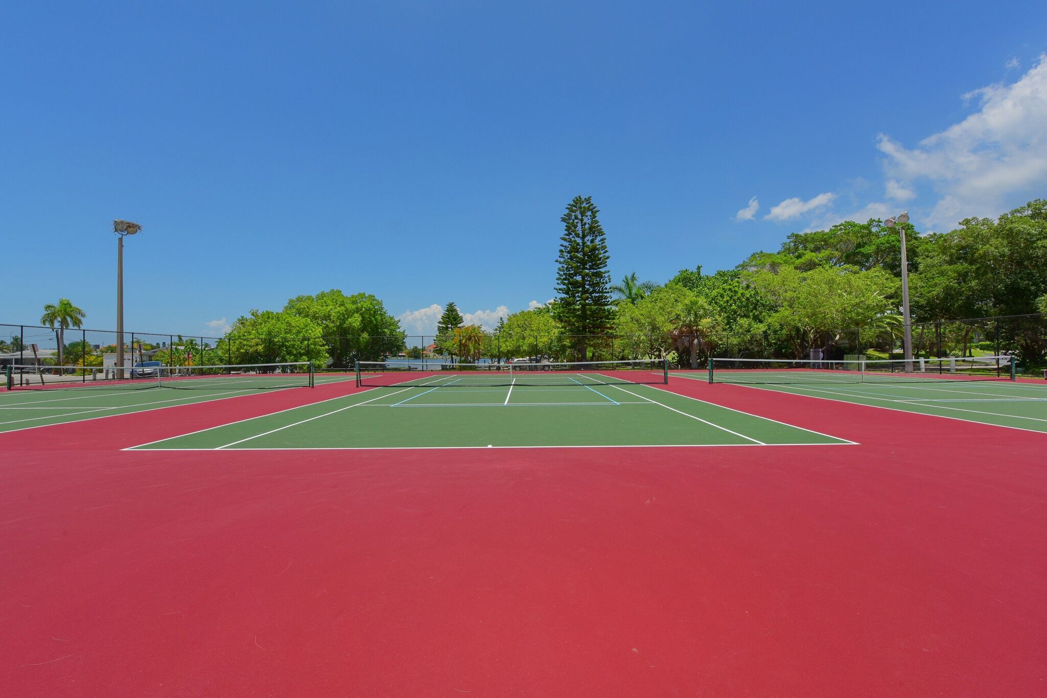 A tennis court with a red and green surface, surrounded by trees and a clear sky.