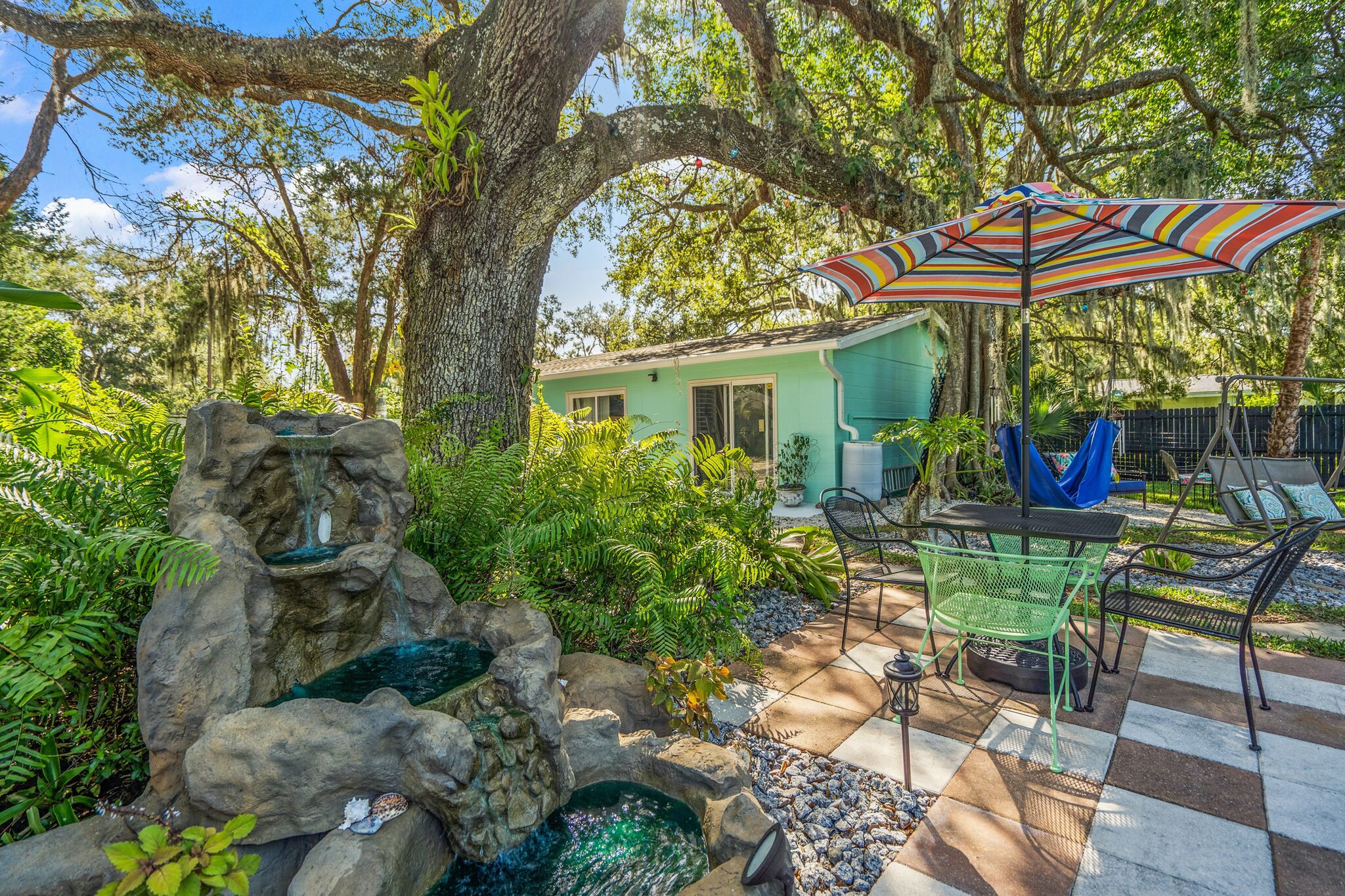 Un espace patio aménagé avec une table et des chaises, un parasol coloré et une petite cascade décorative.