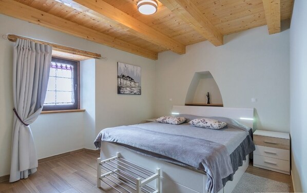 Un dormitorio con techo de madera, una cama con cubrecama gris, una ventana con cortinas, un estante montado en la pared y una mesita de noche pequeña.
