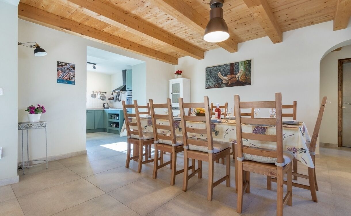 Un coin repas avec des chaises en bois et une table, une cuisine à l’arrière-plan et un plafond avec des poutres apparentes.