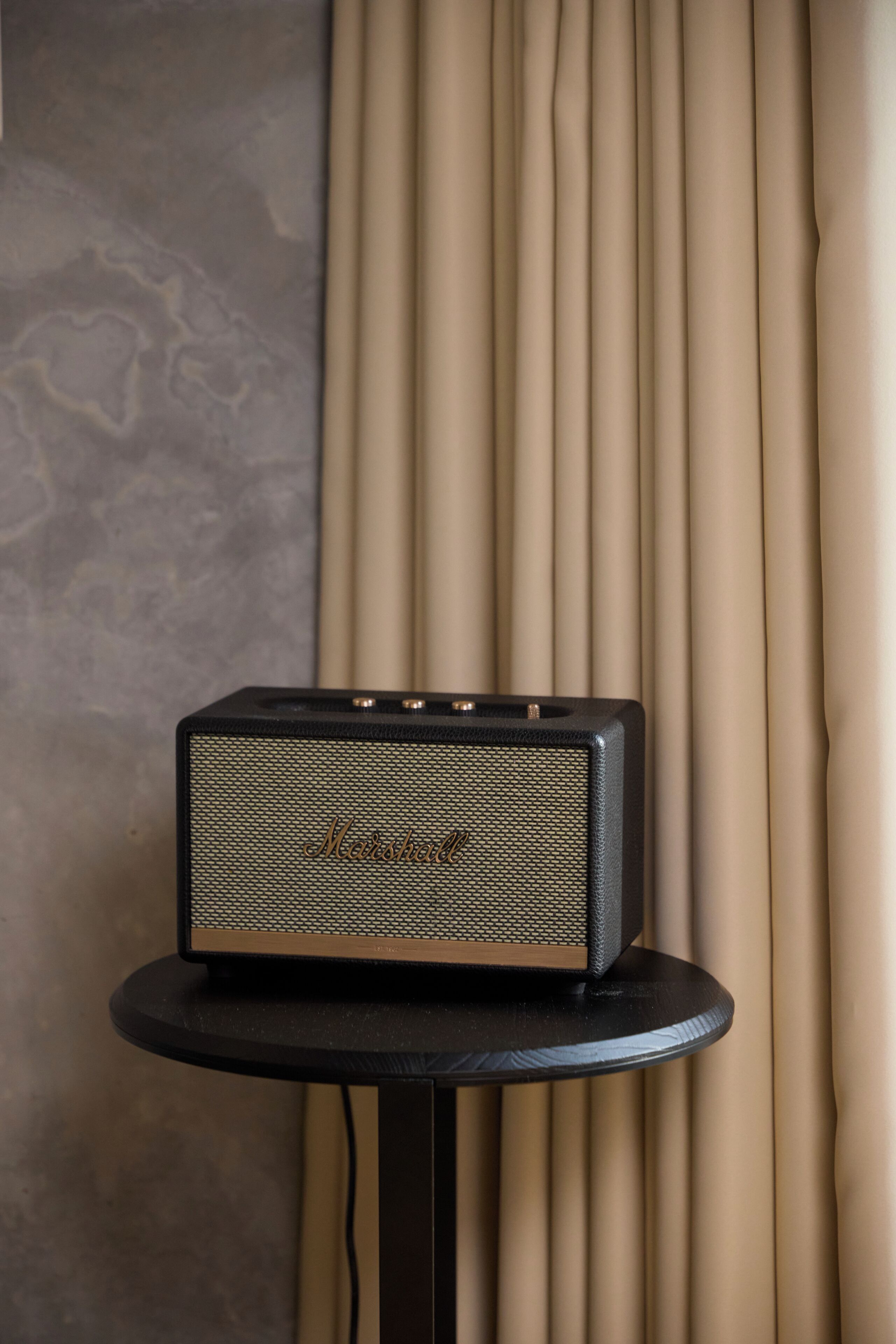 A Marshalls speaker on a small table against a backdrop of beige curtains and a textured wall.