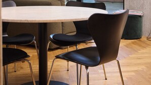 Un comedor moderno con una mesa redonda, sillas negras, una lámpara colgante y vistas a un edificio de ladrillo a través de grandes ventanales.