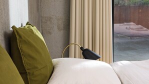 Una cama con almohadas blancas y verdes, una lámpara negra y una ventana con cortinas color beige.