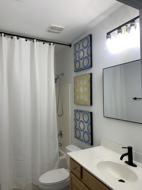 A bathroom with a white toilet, a sink with a black faucet, a shower with a white curtain, and three patterned wall art pieces. - King bed and 4 twins in a 3 BR 2.5 bath Sleeps 6 Heart of Downtown Raleigh (Raleigh)