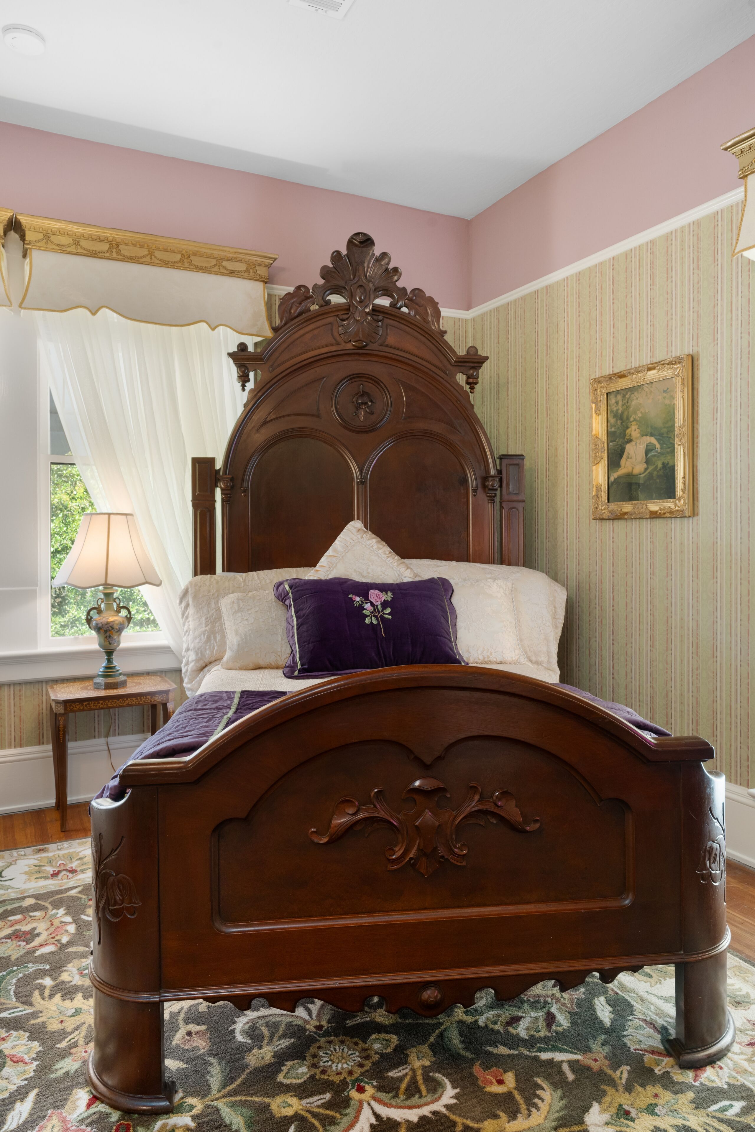 A bedroom with a wooden bed, a nightstand with a lamp, a window with curtains, and a framed picture on the wall.