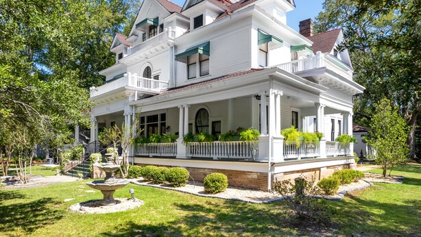 Une grande maison blanche à deux étages, dotée d’un porche à l’avant, de nombreuses fenêtres et d’un jardin bien entretenu avec une fontaine.