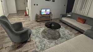A modern living room with a sofa, armchair, and a coffee table. A television is on a stand, and there is a door leading to another room. - Konak Suite Ankara (Ankara)
