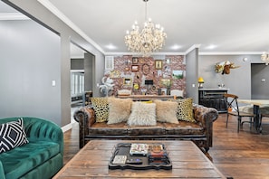 A living room with a green sofa, a brown leather couch with decorative pillows, a wooden coffee table with items, and a brick-patterned wall with framed pictures.