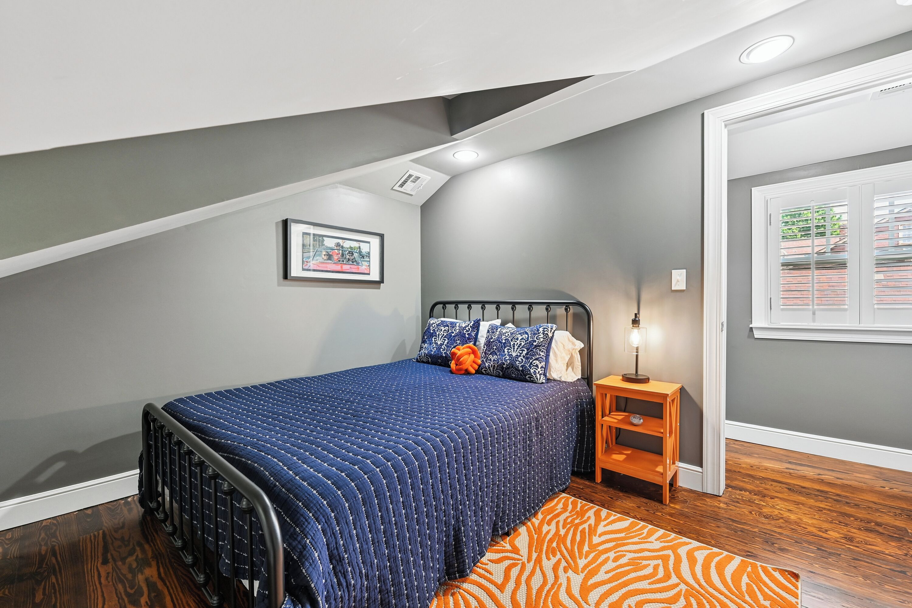 A bedroom with a sloped ceiling, a bed with a blue quilt, a nightstand, a lamp, and a window with blinds.