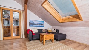 A cozy loft bedroom with a sloped ceiling, a large window, a wooden floor, a sofa, a small table, and a painting of a mountain landscape. - Neu Renovierte Ferienwohnung in Zentraler Ruhiger Lage (Lenggries)