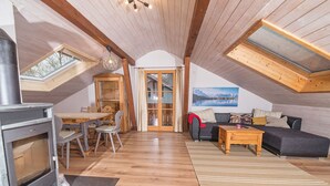 A cozy attic room with a wooden floor, a dining table with chairs, a sofa, a fireplace, and a large window with a view of mountains. - Neu Renovierte Ferienwohnung in Zentraler Ruhiger Lage (Lenggries)