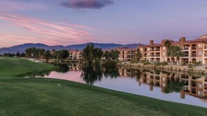 A golf course with a calm lake reflecting buildings, surrounded by greenery and mountains in the background. - Excellent retreat near Coachella Valley festival (Palm Desert)