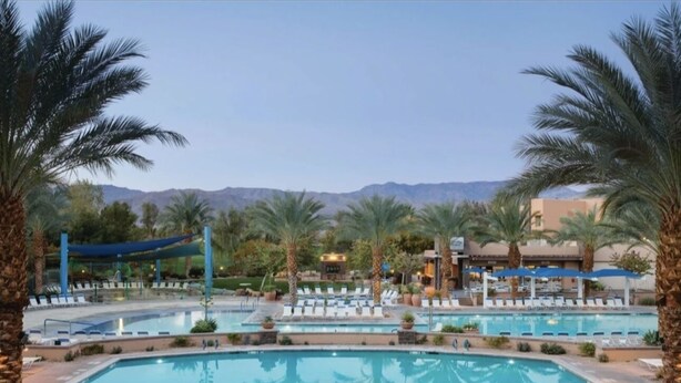 A swimming pool with lounge chairs and palm trees, surrounded by buildings and mountains in the background. - Excellent retreat near Coachella Valley festival (Palm Desert)