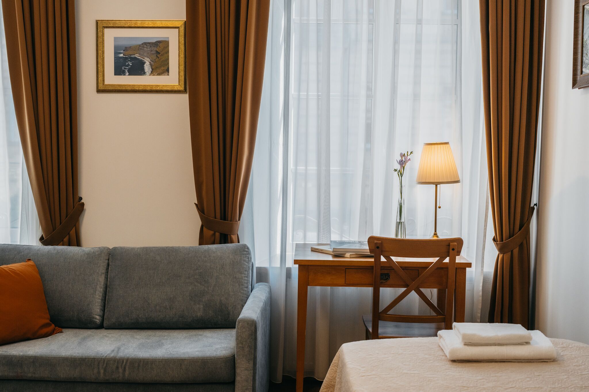 Ett rum med en grå soffa, ett skrivbord i trä med en lampa, en inramad tavla på väggen och en säng med hopvikta handdukar.