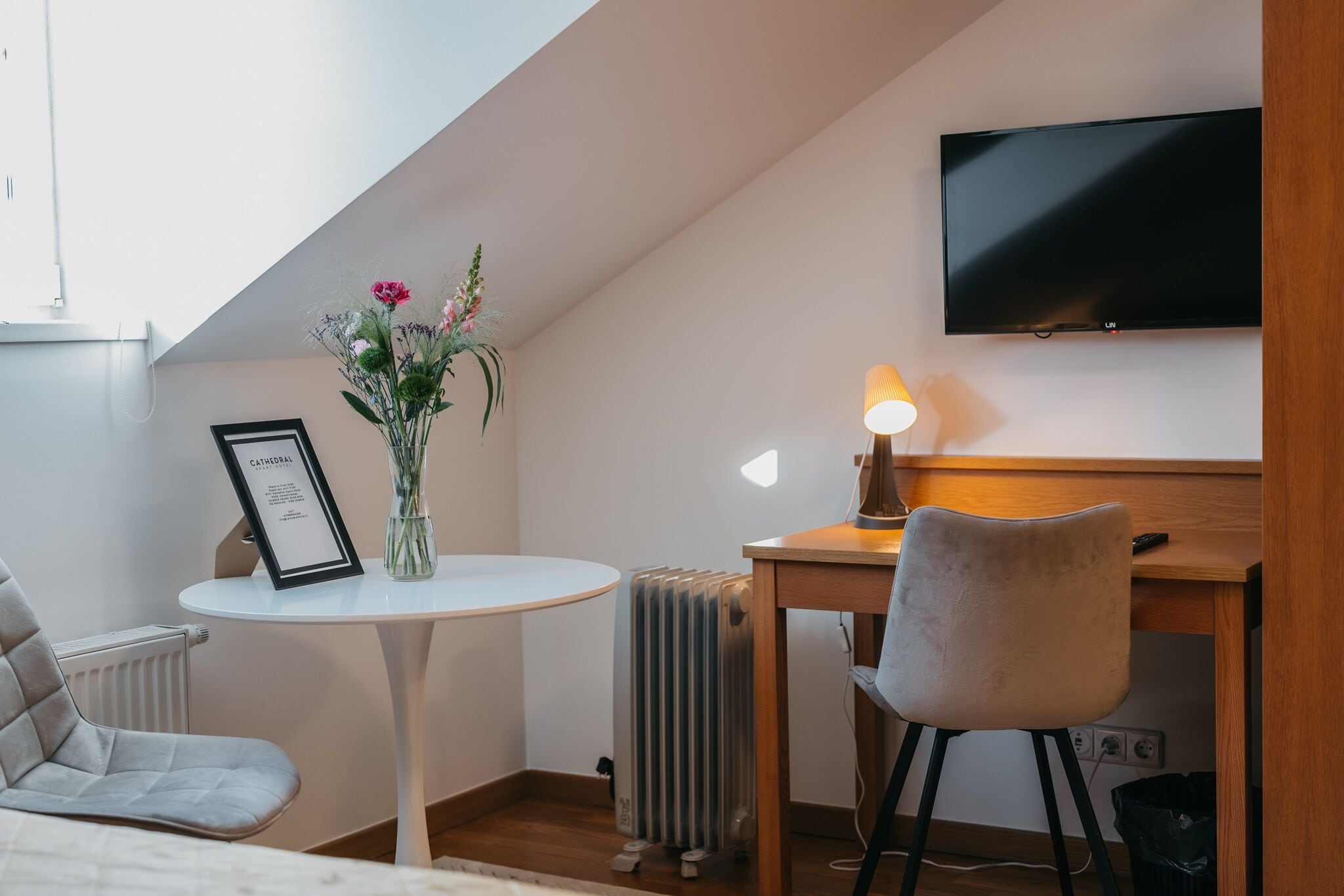 Une petite pièce agréable, dotée d’une table ronde, d’une chaise, d’un radiateur et d’un téléviseur fixé au mur.