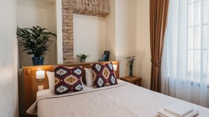 Una cama bien hecha con almohadones estampados, una lámpara de noche, una planta pequeña y un cuadro en una mesita de madera.