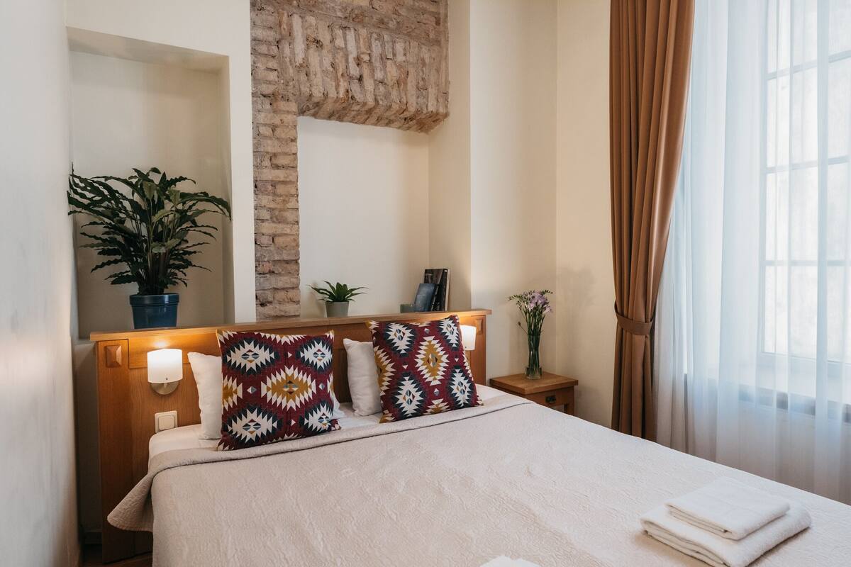 A neatly made bed with patterned pillows, a bedside lamp, a small plant, and a framed picture on a wooden nightstand.