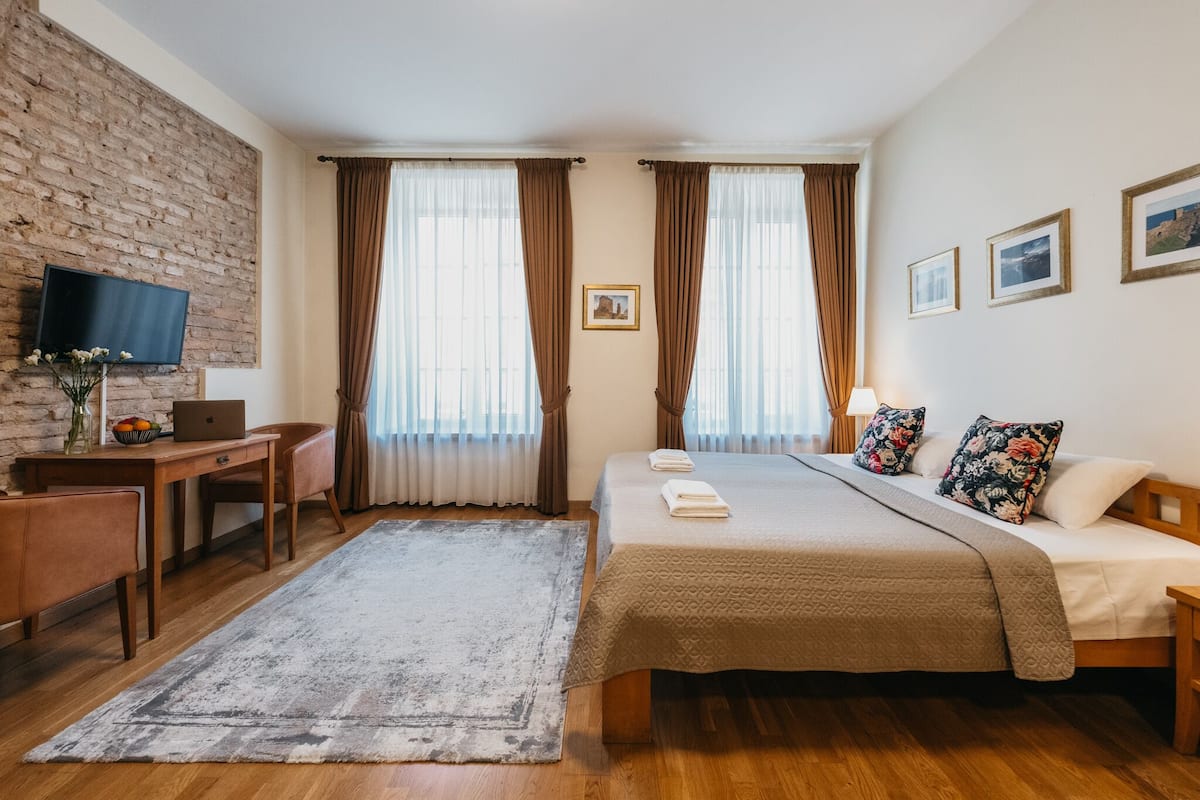 A hotel room with a brick wall, a desk with a laptop, a bed with a beige bedspread, and two windows with brown curtains.