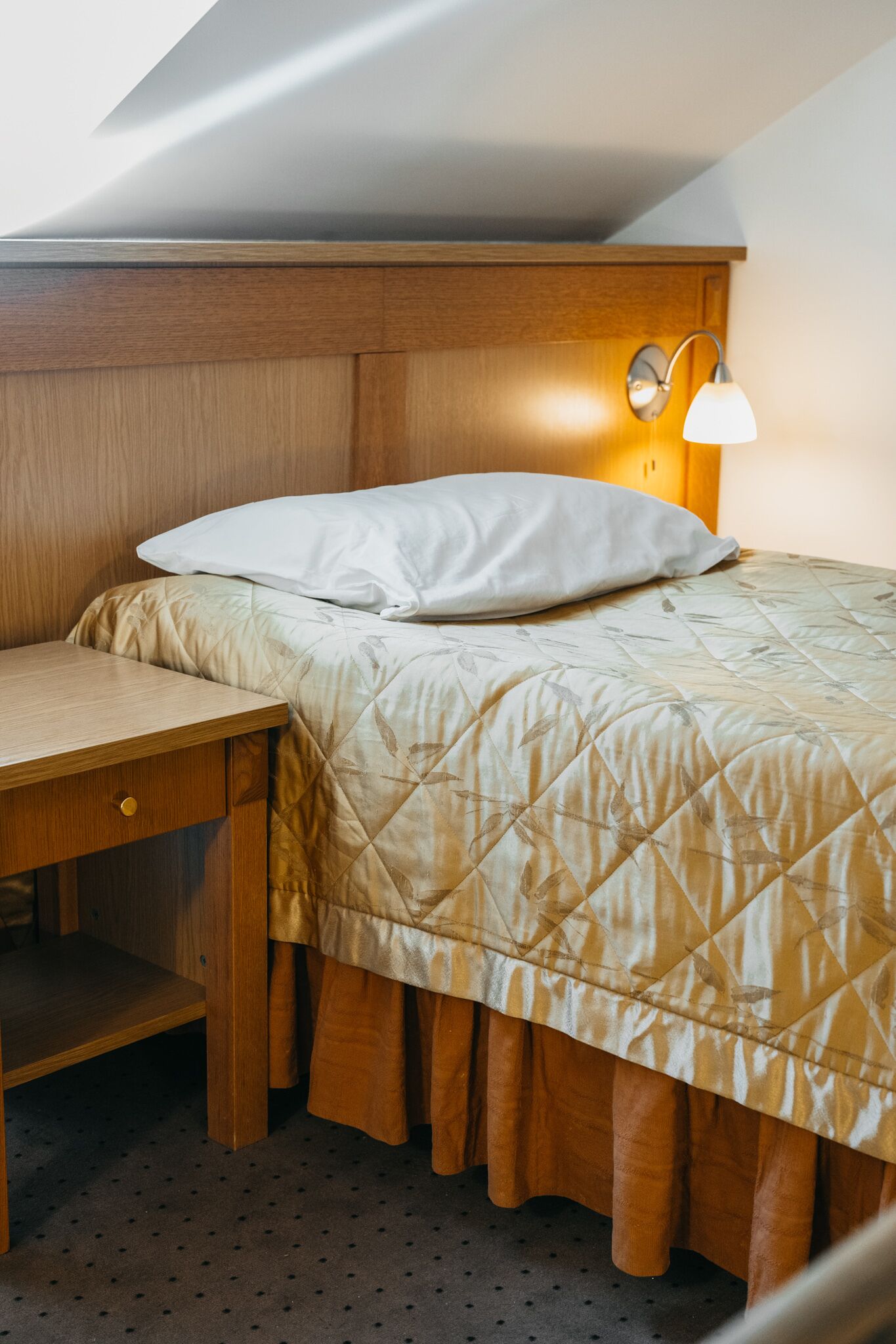 Una cama bien hecha con un edredón acolchado, una mesita de noche con cajón y una lámpara de pared.