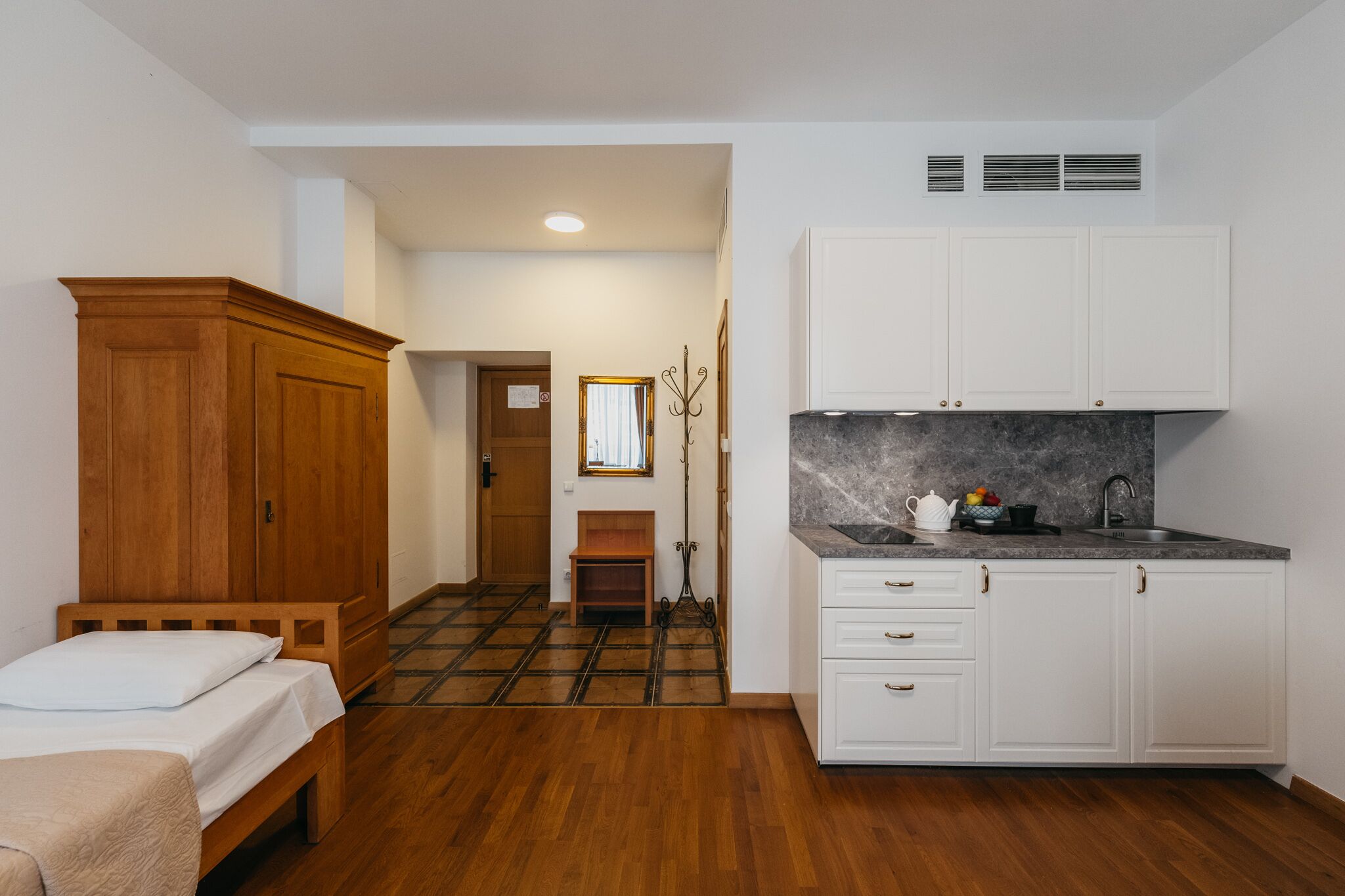Une pièce de taille modeste comprenant un lit en bois, un petit banc, une kitchenette avec des meubles blancs et un plan de travail en marbre, ainsi qu’une armoire en bois.