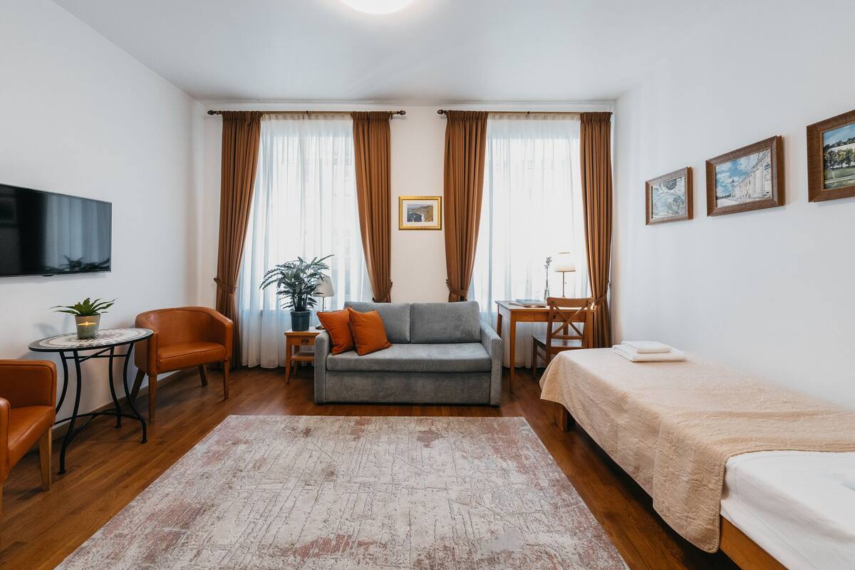 A compact hotel room with a sofa, a bed, a small table with a lamp, a TV, and a potted plant.