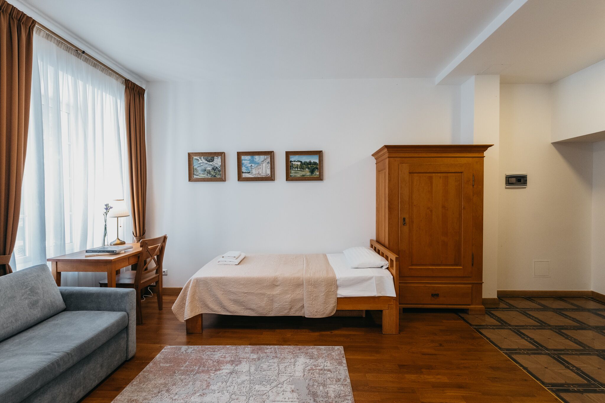 Una habitación pequeña y ordenada con una cama, un armario de madera, un sofá gris, una mesita con una lámpara y tres cuadros enmarcados en la pared.