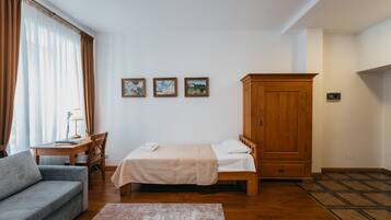 Una habitación pequeña y ordenada con una cama, un armario de madera, un sofá gris, una mesita con una lámpara y tres cuadros enmarcados en la pared.