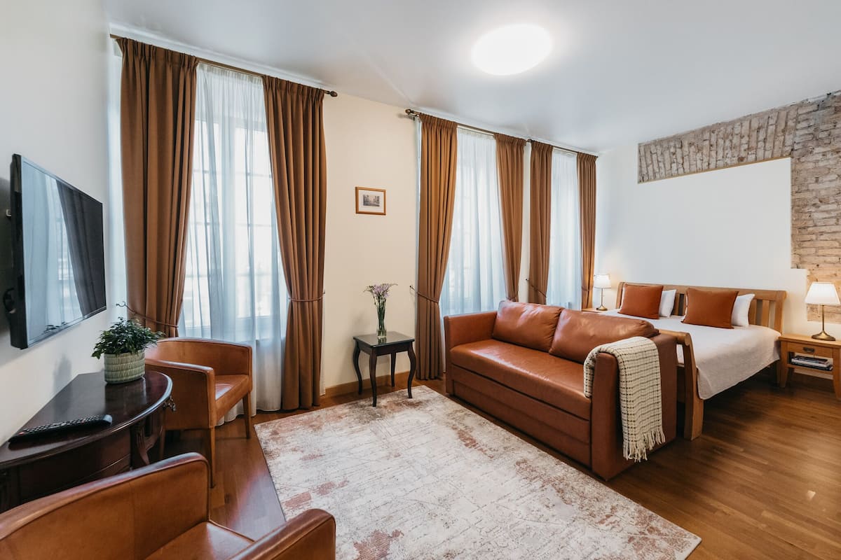 A modern hotel room with a leather sofa, a wooden bed, a small table with a lamp, and a TV on the wall.