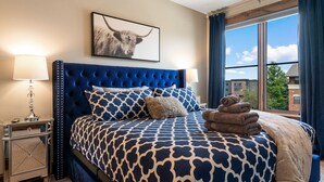 Un dormitorio con cabecera capitonada, ropa de cama estampada, una lámpara de noche y vistas a edificios a través de una ventana.