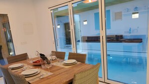 Ein Essbereich mit einem Holztisch, gedeckt für sechs Personen, umgeben von Korbstühlen, mit einer Glastrennwand und Blick auf ein Schwimmbad und eine Außenlounge.
