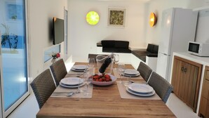 Ein moderner Essbereich mit einem Holztisch, gedeckt für vier Personen, umgeben von Korbstühlen. Auf dem Tisch steht eine Obstschale, und der Raum verfügt über ein großes Fenster, einen Fernseher und einen Kühlschrank.