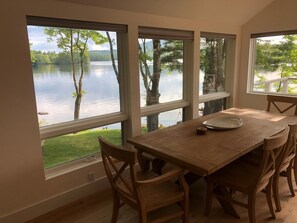 Uma área de refeições com mesa e cadeiras de madeira, janelas amplas com vista para um lago e um corrimão branco na parte externa.