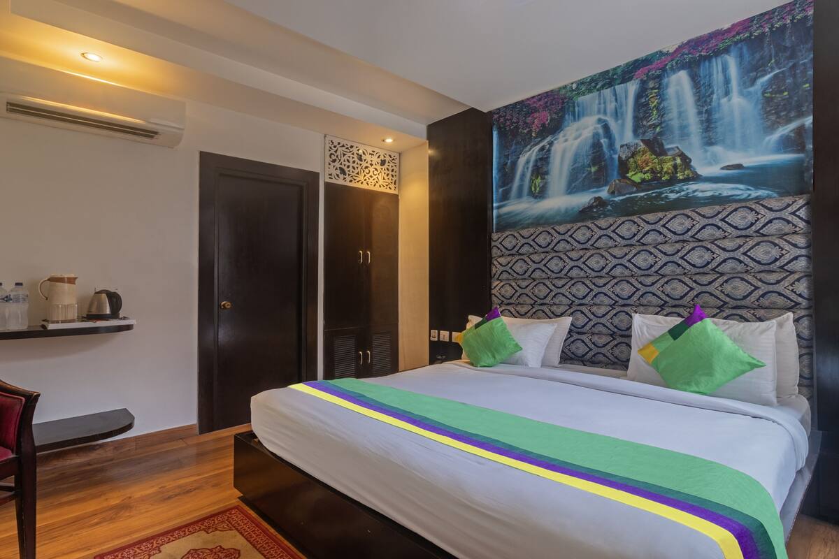 A hotel room with a bed, a chair, a desk, a wardrobe, and a wall with a waterfall painting.