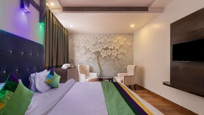 A modern hotel room with a large bed, two white armchairs, a small round table, and a flat-screen TV mounted on the wall. - Treebo The Royal Taj (Agra)