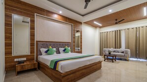 A modern bedroom with a wooden headboard, a large bed with colorful pillows, a bedside table with a phone, a sofa, and a ceiling fan. - Treebo Omnibliss (Dera Bassi)
