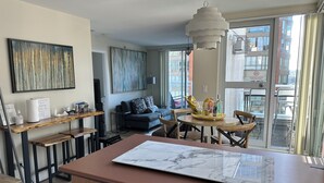 A modern living room with a dining area, a sofa, a coffee table, and a large window with a view of buildings. - Penthouse with Large Deck on Seawall / Beach w/ Views (Vancouver)
