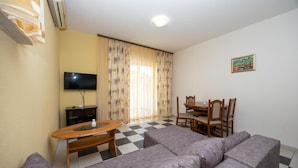 Ein Wohnzimmer mit einer Sofagarnitur, einem Esstisch mit Stühlen, einem Fernseher und einem gemusterten Vorhang.
