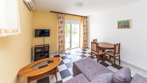 Ein Wohnzimmer mit einer Couch, einem Couchtisch aus Holz, einem Fernseher, einem Esstisch mit Stühlen und einer Glasschiebetür zu einem Außenbereich.