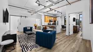 Una sala de estar moderna con un sofá, una mesa de centro y un televisor. Hay una zona de cocina con una mesa de comedor y sillas. El cuarto tiene piso de madera y paredes blancas.