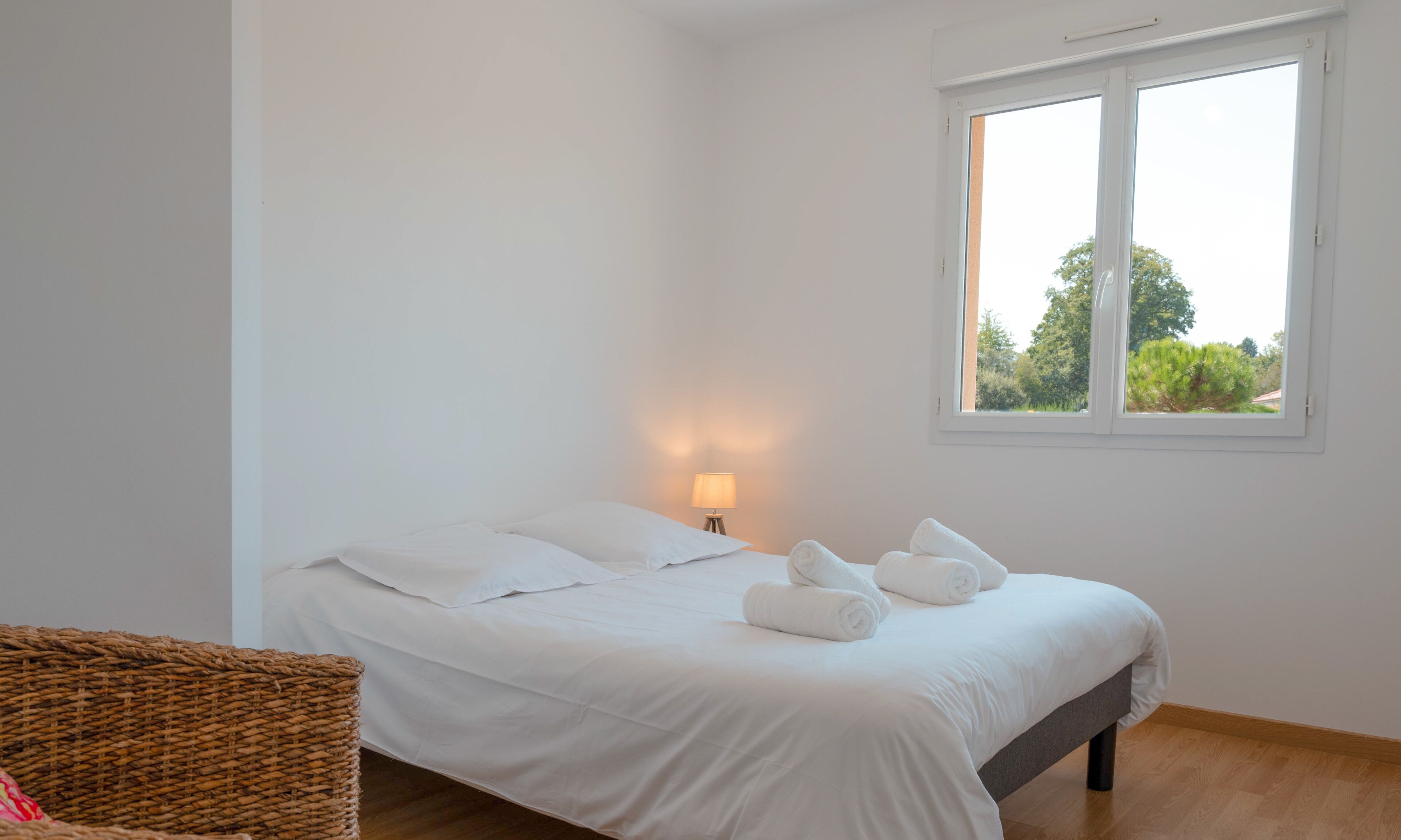 A neatly made bed with white linens and rolled towels, a bedside lamp, and a window with a view of greenery.