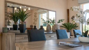 Un comedor con una mesa de madera, sillas y un espejo grande. Hay plantas en macetas y un jarrón con flores sobre la mesa.